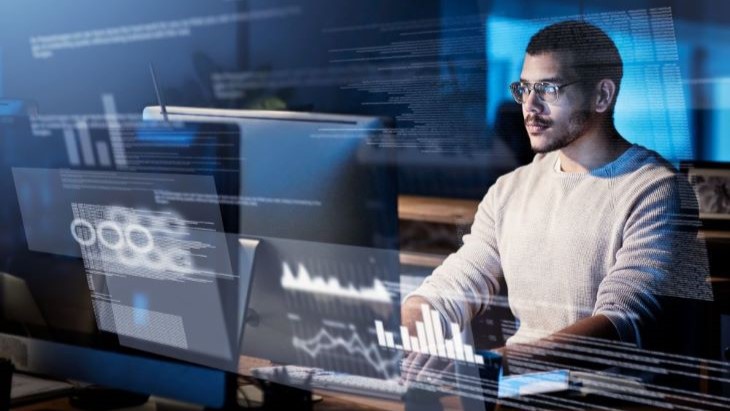 Alt - A person working at a computer with multiple screens displaying various graphs and data visualisations. The setting appears to be a modern tech workspace, emphasising data analysis or software development.