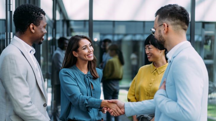 Alt - The image depicts a modern office setting where four individuals are standing and engaging in conversation. Two of them are shaking hands, suggesting a greeting or agreement. One person is dressed in a light grey suit, while the others wear a mix of professional attire, including a blue blouse and a yellow blouse. The background features glass walls and reflections, contributing to the sleek, contemporary atmosphere of the office. Additional people are visible, adding to the dynamic and busy feel of the environment.