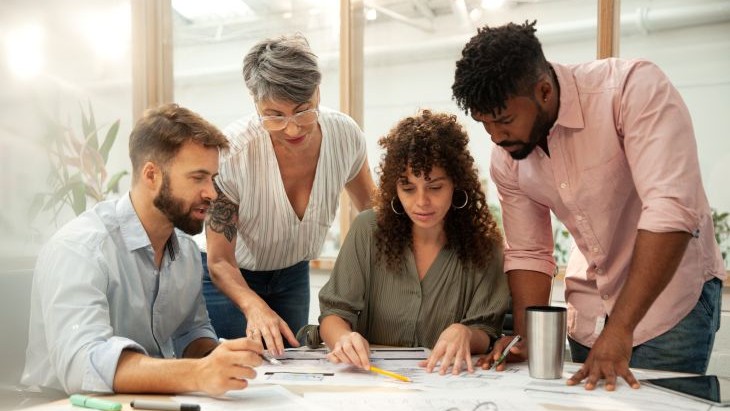 Alt - Four individuals are gathered around a table in a modern office setting, actively engaged in reviewing documents and sketches. The scene conveys a collaborative work session, with glass partitions and indoor plants in the background adding to the professional and creative atmosphere.
