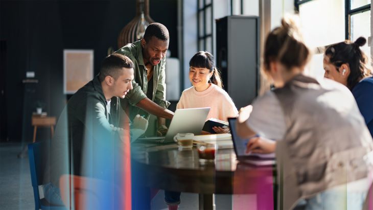 Alt- A group of people are gathered around a table in a modern, well-lit room with large windows. They are engaged in a collaborative discussion, with one person standing and pointing at a laptop screen while others are seated, looking at devices and papers. The setting suggests a casual yet productive work environment, possibly a brainstorming session or team meeting. The image highlights teamwork, technology, and a shared workspace.