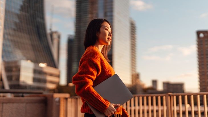 Alt - Person in a bright orange sweater holding a laptop, standing outdoors during golden hour in a modern cityscape with glass high-rise buildings in the background.