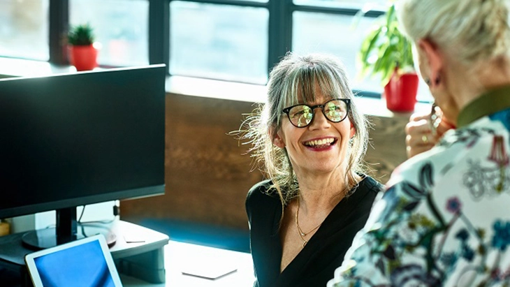 Woman seated at workstation looking up and smiling at another woman