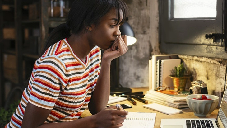 Young woman sitting at desk, looking at laptop screen and taking written notes