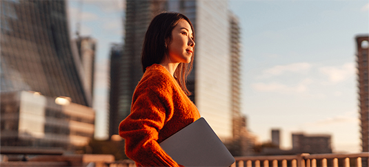 Person wearing an orange sweater holding a closed laptop while standing outdoors on a balcony with modern skyscrapers in the background during sunset