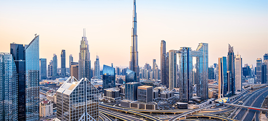 Panoramic view of Dubai city skyline featuring modern skyscrapers, including the Burj Khalifa, with highways and overpasses in the foreground under a clear sky