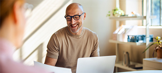 Person seated at a desk in a bright office environment, working on a laptop and holding documents, with shelves and plants in the background.